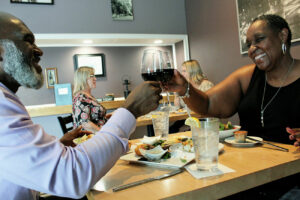 A couple sits at a table with food in front of them, clinking their wine glasses together.