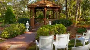A garden with red brick paths, a wooden gazebo and white chairs for sitting on the grassy lawn besides it. The garden is filled with green trees and shrubs.