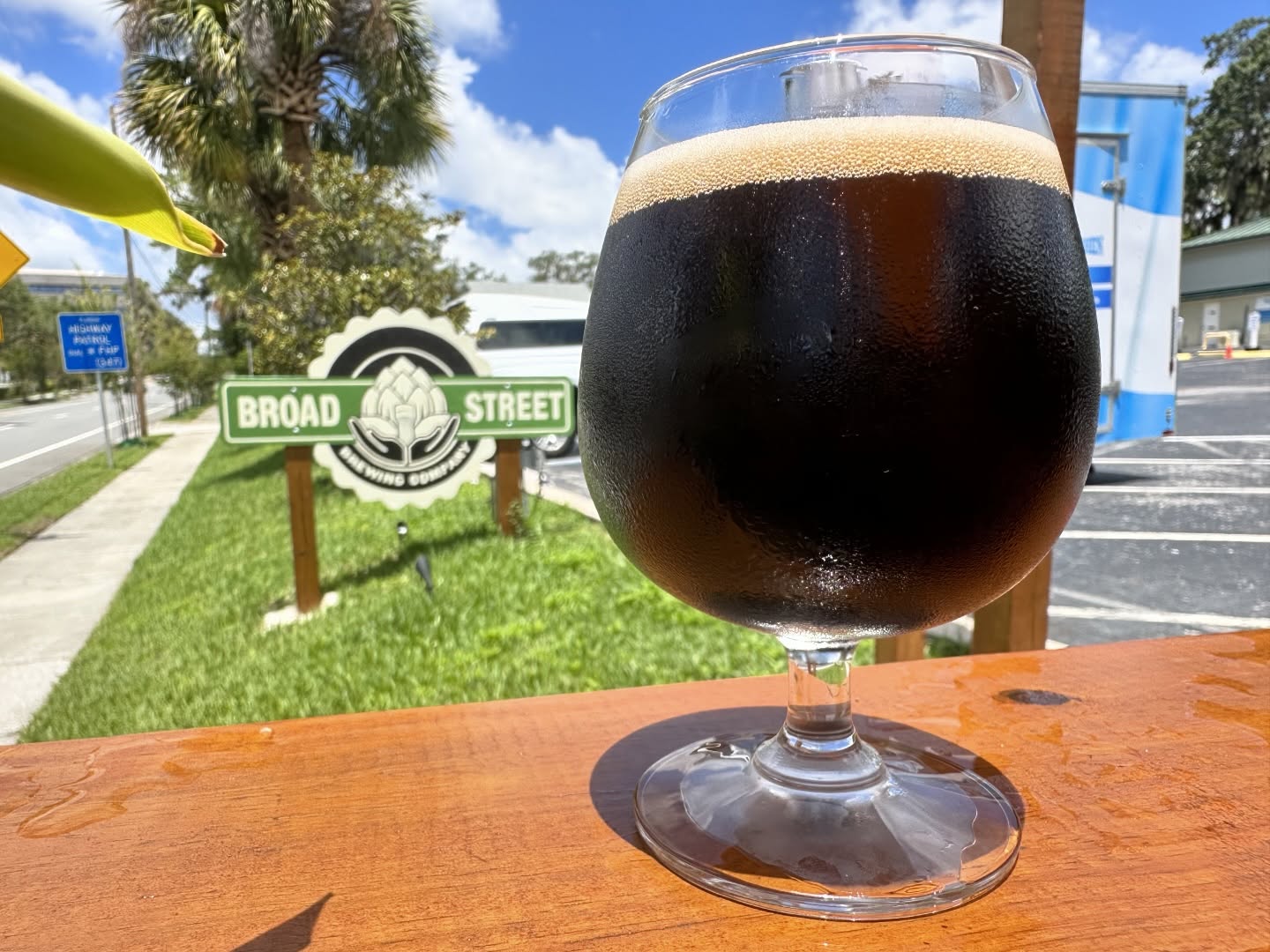 Close up of a dark colored beer on a wooden counter. In the background are trees, green grass, and a sign for 'Broad Street Brewing' featuring the Brooksville Water Tower