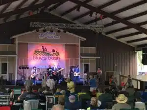 A audience with seated people in an open-air pavilion watches a band perform. The band has men with guitars and a drum set, standing on a wooden platform with a banner behind that reads 'Blues Bash.'