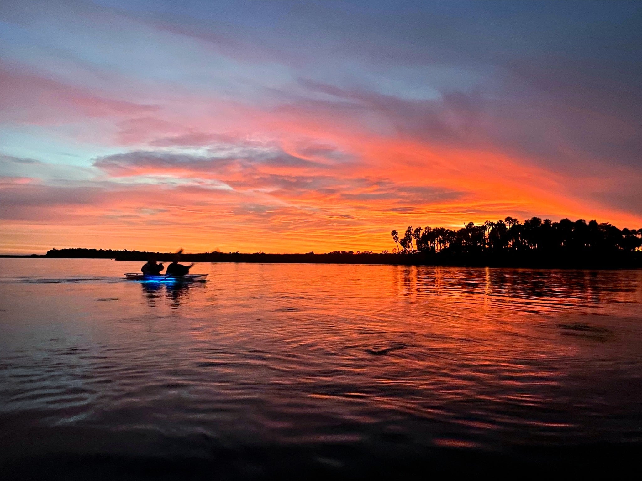 Two people in a clear kayak paddling on tranquil coastal waters reflecting the sunset with bright orange and pink clouds in the sky.