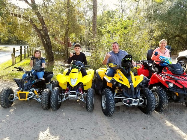 A group four with two adults and two kids each sit on their own ATV, smiling and giving the 'gnarly' hand sign.