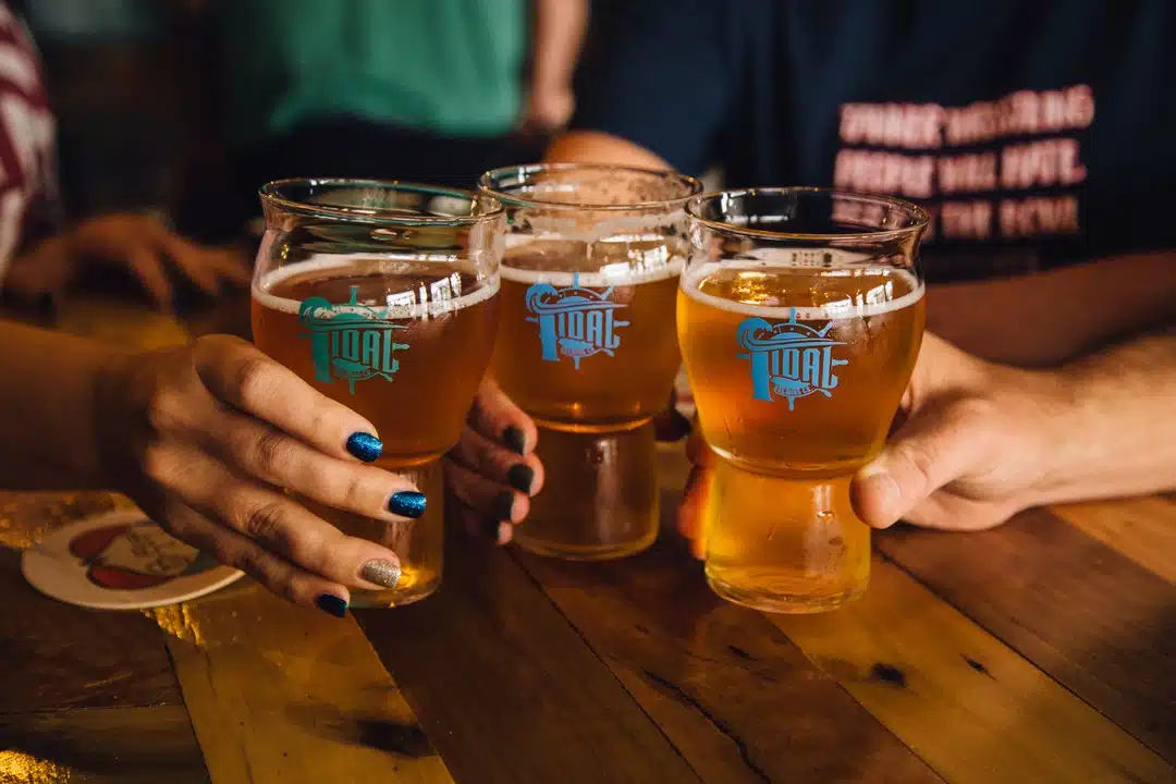Three hands, each holding a glass of beer, come together for a toast on a wooden table. The cups each have a blue logo for 'Tdial Brewing Co." on them.