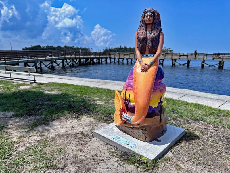 A mermaid statue with brown skin and hair, and an orange mermaid tail, part of The Mermaid Tale Trail. The statue is standing in front of the fishing pier at Bayport Park.