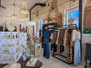 A woman shops while browsing items on a clothing rack, with displays on jewelry on a table nearby. The walls of the shop are white white with red brick in places, and chandeliers hang from the ceiling.