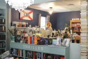 Inside a book store, where a low shelf filled with books has a sign "THRILLER" and a coffee shop behind the bookshelf. A wooden board with the Florida flag is on the wall behind it.