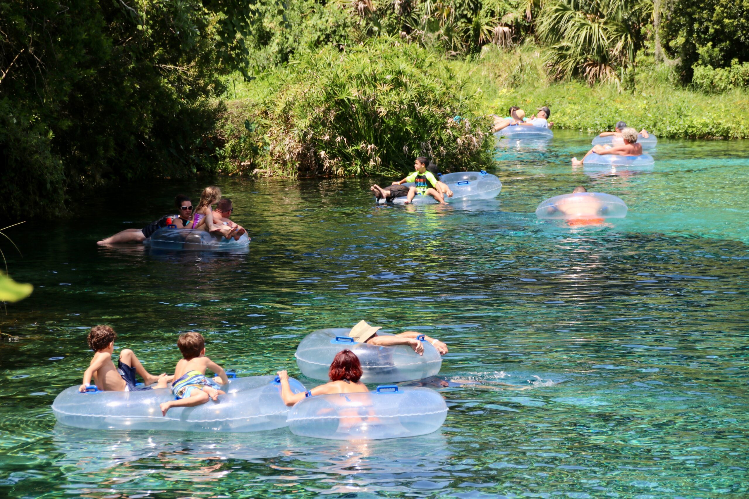 A group of people floating on turquoise water in clear inflatable inner tubes. Lush foliage and trees shade the riverside.