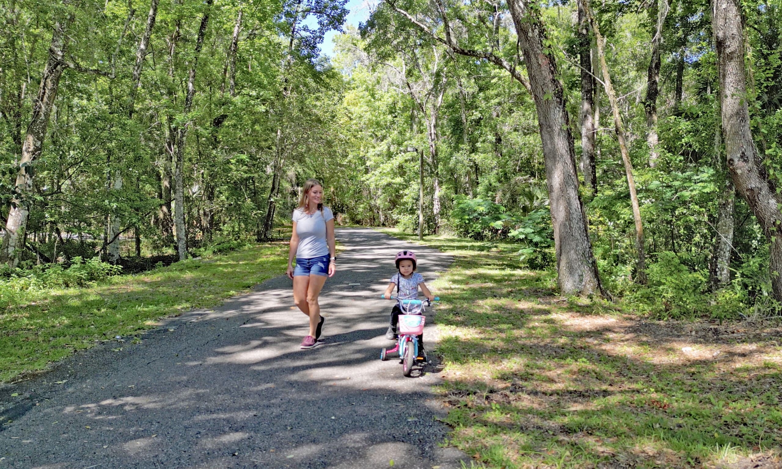 A woman walks along a shaded paved path next to a small girl on a bicycle. The paved trail is lined by lush trees and greenery.
