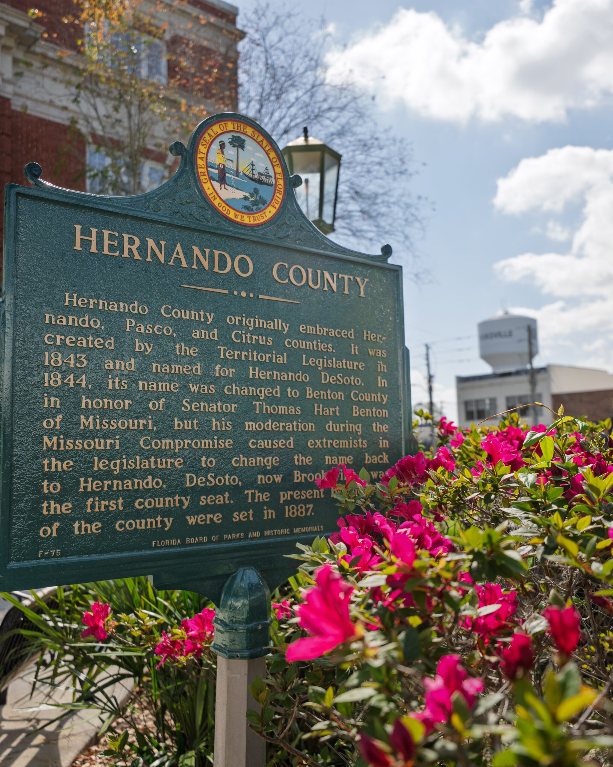 Historic Hernando County Courthouse Marker A dark green sign with text describing the history of Hernando County in front of a red brick courthouse. A bush with azaleas blooms in front of the sign.