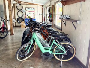 A row of bicycles (e-bikes) stationed inside of a building. The adjacent wall has a shelf with bike helmets, and a tool box and bike tires can be seen in the back of the room.